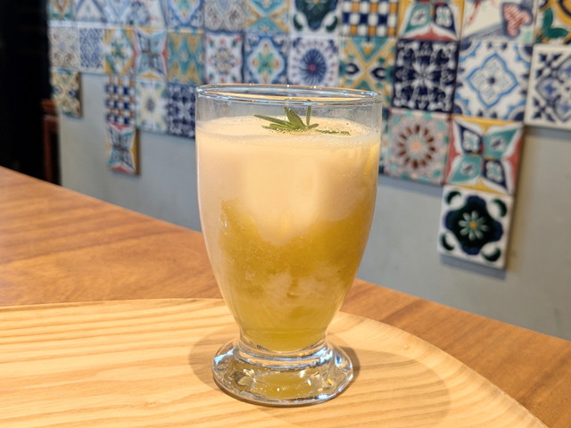 グラスの底に黄色いパイナップルソース、上にアーモンドミルクのフォームが重なりローズマリーが添えられたドリンク