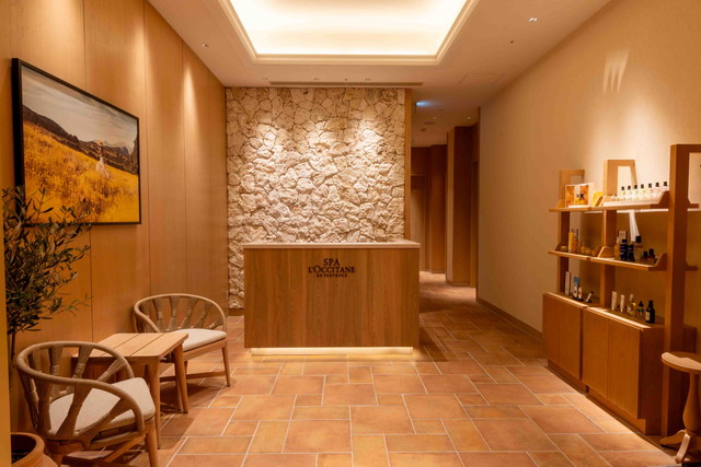 SPA L'OCCITANE EN PROVENCEのエントランス・受付。石壁にロゴ、オリーブツリーと木の温もりある空間